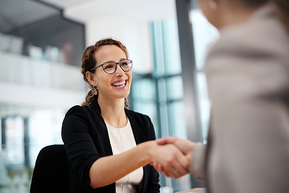 A young woman wearing glasses is shaking hands with another woman and smiling.